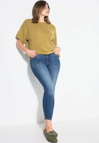 Hořčicový pletený svetr s krátkým rukávem, kombinovaný s těsně přiléhajícími modrými džíny a olivově zelenými slip-on botami se stříbrným řetízkem.