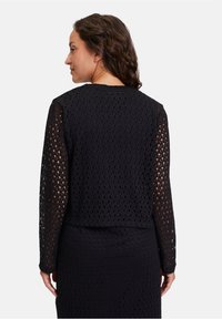 Schwarze Langarm-Strickjacke mit einem diamantförmigen, offenen Strickdesign. Der Rücken ist leicht verkürzt mit einem runden Halsausschnitt und gerippten Bündchen.