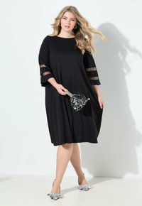 Eine Frau in einem schwarzen Kleid mit transparenten Ärmeln hält eine glitzernde Clutch und steht vor einem hellen Hintergrund, wobei sie elegantes Schuhwerk präsentiert.