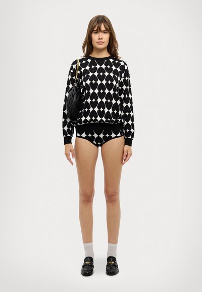 Sweater patterned em preto e branco com formas de coração, combinado com roupa interior correspondente. Modelo segura uma bolsa preta e usa sapatos pretos com meias.