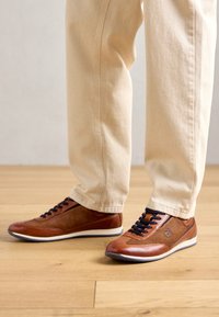 Baskets en cuir marron avec des lacets noirs portées avec un pantalon beige sur un sol en bois clair, jambes visibles à partir du milieu du mollet.
