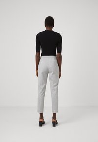 Osoba stojąca tyłem, ubrana w czarną obcisłą bluzkę, szare dopasowane spodnie długości do kostek oraz czarne buty na obcasie, na jednolitym tle.