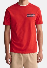 Rotes Baumwoll-T-Shirt mit kurzen Ärmeln und Rundhalsausschnitt, mit einem marineblauen Grafikdruck auf der linken Brust, der "CANT" liest.