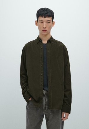 Dunkelgrünes Hemd mit Knöpfen und Kragen, mit weichem Material, langen Ärmeln und lockerer Passform, getragen über einem dunkelgrauen Pullover und Jeans.
