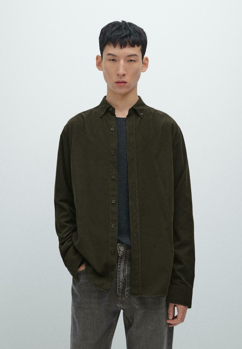 Camicia button-up verde scuro con colletto, caratterizzata da una consistenza morbida, maniche lunghe e una vestibilità comoda, indossata sopra un maglione grigio scuro e jeans.