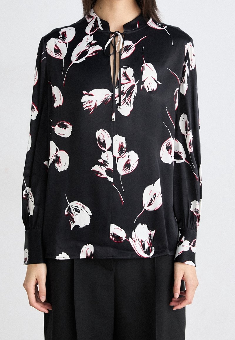 Blouse noire avec motif floral blanc et rouge, col montant, détail noué au niveau de l'encolure, et manches longues avec poignets froncés.
