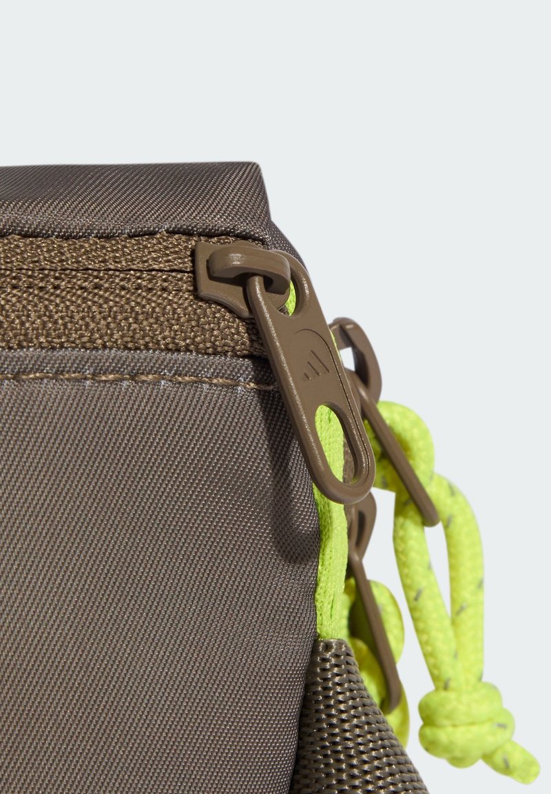 Braune Stofftaschenecke mit braunem Reißverschluss und neongelben Kordeln, die durch die Reißverschlusszüge gezogen sind, vor hellem Hintergrund.