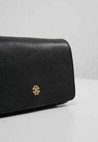 Černá kožená peněženka s texturovaným povrchem, na přední straně zdobená zlatou ozdobou ve tvaru květiny a kontrastním šitím.