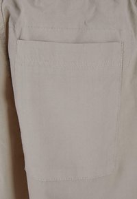 Beige stof med en glat overflade, der har en flad, stitched lomme. Designet inkluderer rene linjer og minimale accenter.