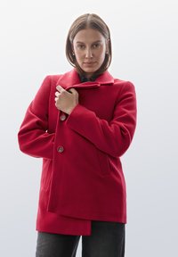 Abrigo de lana rojo con un diseño de doble botonadura, gran collar y detalles de botones. El modelo sostiene el collar con una camisa negra debajo.