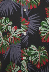 Tela negra con hojas de monstera en verde, blanco y rojo, y frondas de palma moradas, que presenta una fila vertical de botones beige en el centro.