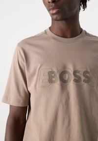 Béžové tričko s krátkým rukávem vyrobené z měkké bavlny, s vyraženým logem "BOSS" v různých texturách na hrudi.