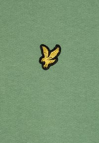 Tessuto lavorato a maglia verde con un uccello ricamato giallo su un contorno nero, con una texture liscia e un design casual e minimalista.