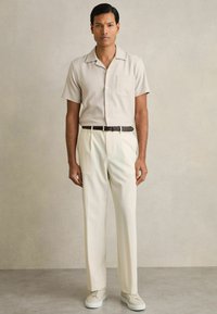 Cămașă scurtă bej deschis, cu un buzunar, asociată cu pantaloni crem tailor-made și adidași de culoare deschisă. Design simplu și curat.