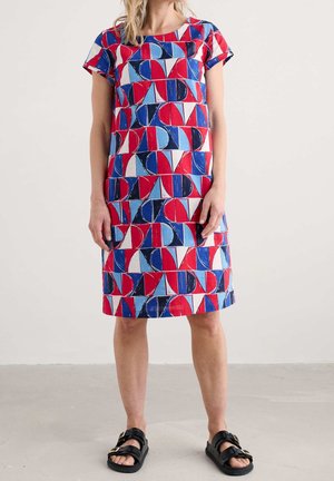 Femme portant une robe mi-longue à manches courtes, avec un motif géométrique rouge, bleu et blanc, associée à des sandales noires à boucle.