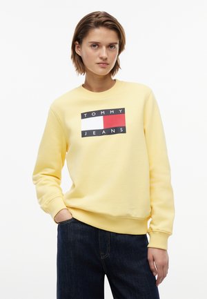 Młoda osoba z krótkimi włosami, ubrana w żółtą bluzę Tommy Jeans oraz ciemne dżinsy, stoi z jedną ręką w kieszeni.