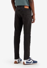 Svart denimjeans med rak benform, fem fickor och en brun läderlapp på baksidan. Bärs med blå och vita träningsskor.