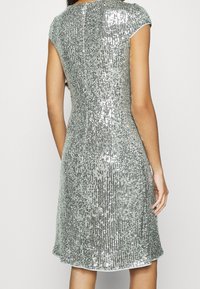 Robe en sequins argentés avec une coupe ajustée et des manches courtes. Texture scintillante se déplaçant verticalement, avec une fermeture éclair dissimulée dans le dos.