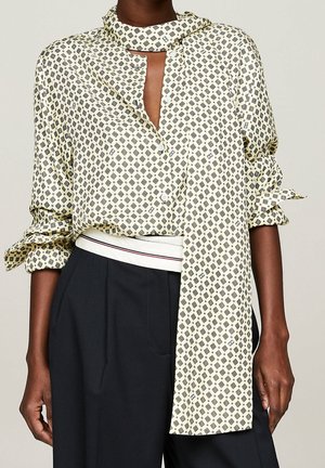 Person trägt eine gemusterte cremefarbene Bluse mit schalähnlichem Detail und hochgekrempelten Ärmeln, kombiniert mit schwarzen, hoch taillierten Hosen.