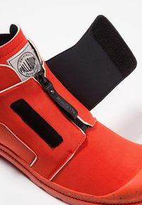 Chaussure montante orange vif avec fermeture éclair noire et bride Velcro, languette de tirage de marque, et écusson blanc avec logo sur le col de la cheville.