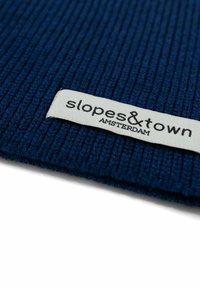 Tessuto a coste navy con etichetta bianca che riporta la scritta "slopes&town Amsterdam", caratterizzata da un testo nero in un font moderno.