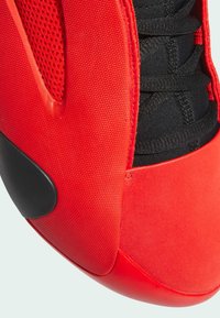 Zapato deportivo rojo con parte superior de malla texturizada, puntera de goma lisa y sistema de cordones de tela negra. Diseño curvado con talón redondeado.