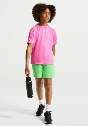 Kind mit lockigem Haar läuft und hält eine schwarze Wasserflasche, trägt ein pinkes Sportshirt, grüne Shorts, weiße Socken und schwarze Turnschuhe.