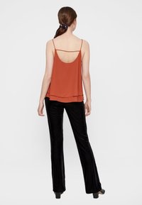 Rdzawoczerwony bezrękawnik z okrągłym dekoltem i warstwowym fasonem; zestawiony z czarnymi, teksturowanymi spodniami w dzwony i czarnymi butami na obcasie.