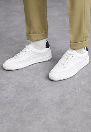 Zapatillas blancas con un acabado de cuero texturizado, punta redondeada y acento negro en el talón. Suela de goma con un pequeño patrón de relieve y detalle de costura.