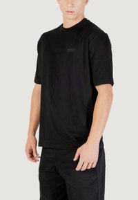 Camiseta de algodón negra con mangas cortas, cuello redondo y una pequeña etiqueta rectangular en el pecho. Diseño simple y de corte relajado.