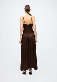 Long brown slip dress with thin straps, fitted bodice, and flowing skirt. Smooth satin texture with a subtle sheen, zips at the back.