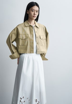 Žena s dlhými čiernymi vlasmi má na sebe béžovú krátku bundu cez biely top a bielu sukňu s kvetinovými výrezmi, stojí na jednoduchom pozadí.