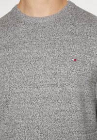 Pull en tricot gris avec un col rond et un tissu texturé. Présente un petit logo en bleu, rouge et blanc sur le côté gauche.