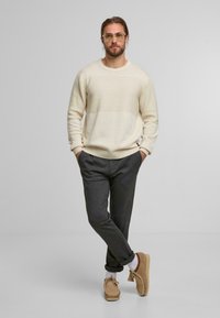 Maglione a maglia crema con motivo testurizzato, pantaloni grigi su misura e pantofole mocassino beige. Il modello è in piedi con le mani in tasca su uno sfondo semplice.