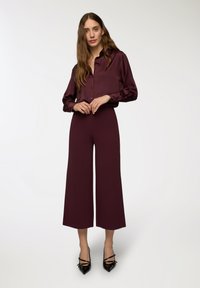 Camicia lunga in satin bordeaux con maniche lunghe abbinata a pantaloni a gamba larga, lunghi fino al pavimento, caratterizzati da una texture liscia e una semplice chiusura con bottone.