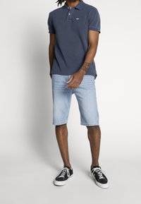 Polo blu navy con un piccolo logo, abbinata a pantaloncini di jeans azzurri e sneakers nere con lacci e suola bianchi.