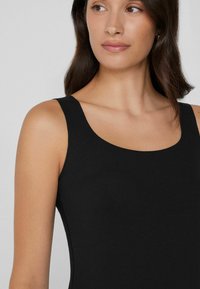 Jonge vrouw met donkerbruin haar, gekleed in een mouwloze zwarte top, die iets naar links kijkt tegen een lichtgrijze achtergrond.
