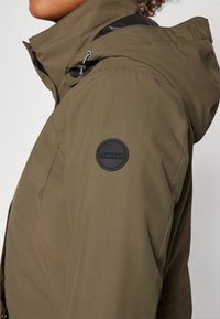 Olivgrön huva jacka med en slät textur, som har en svart cirkulär logotyp patch på ärmen och en kontrasterande axeldesign.