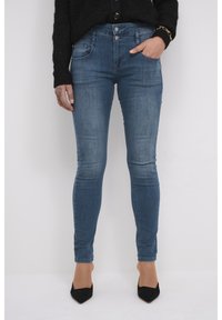 Mršave plave traperice s istrošenim pranjem, izrađene od trapera. Imaju pet džepova, zatvaranje na gumb te uski kroj kroz noge.