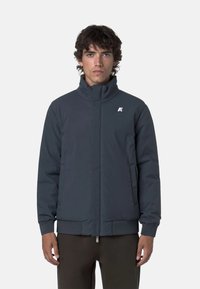 Giacca imbottita da uomo di colore blu navy con colletto alto, zip frontale e tasche laterali. Presenta polsini a coste e un piccolo logo bianco sul petto.