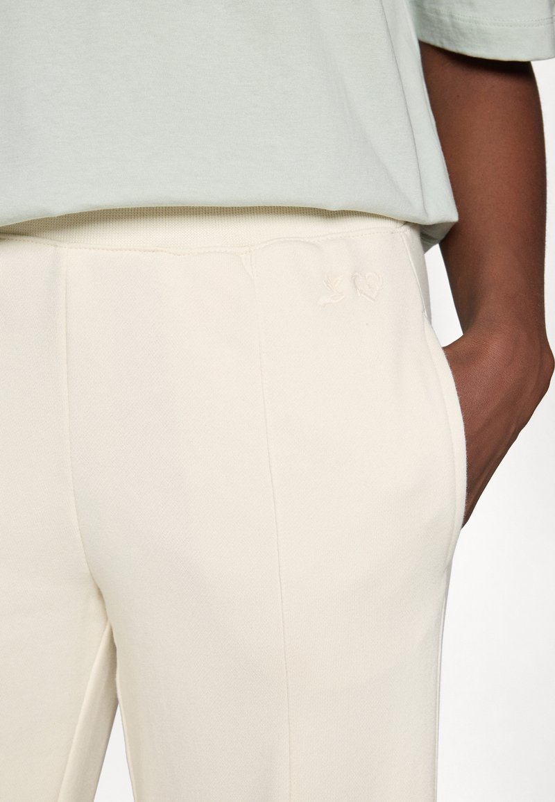 Pantaloni color crema con una texture liscia, caratterizzati da un delicato ricamo di un uccello e un cuore vicino alla tasca anteriore.