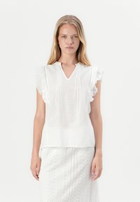 Weiße Bluse mit Rüschensäumen, V-Ausschnitt und strukturiertem Punktmuster. Leichter Stoff, kombiniert mit einem gemusterten weißen Rock.