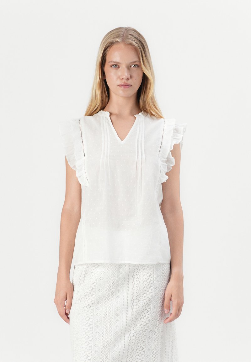 Weiße Bluse mit Rüschensäumen, V-Ausschnitt und strukturiertem Punktmuster. Leichter Stoff, kombiniert mit einem gemusterten weißen Rock.