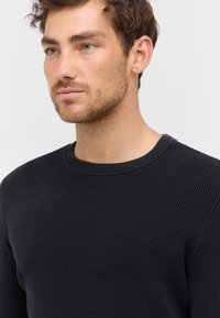 Pull en tricot noir avec un col rond. Présente un tissage texturé et des poignets côtelés. Coupe simple et ajustée sans embellissements supplémentaires.