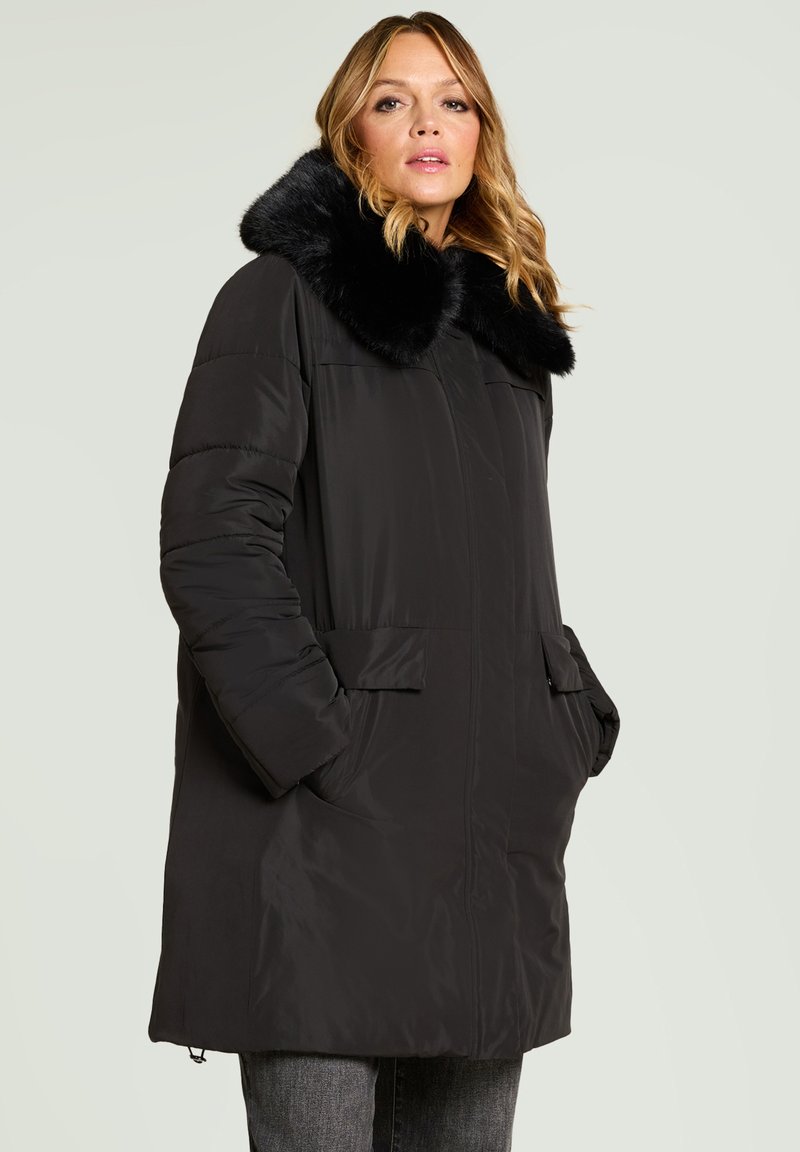 Abrigo negro acolchado con cuello de piel sintética, presenta cierre frontal con cremallera, dos bolsillos laterales y un corte holgado. Textura suave de nylon, diseño hasta la rodilla.