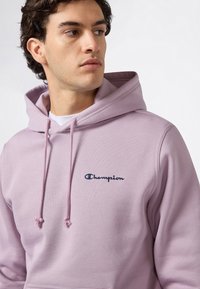 Młody mężczyzna w jasnofioletowym hoodie Champion z regulowanym kapturem, patrzący w bok na jednolitym białym tle.