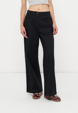 Femme portant un pantalon noir taille haute à jambes larges, un top court gris clair et des chaussures plates rouge foncé, debout devant un fond blanc.