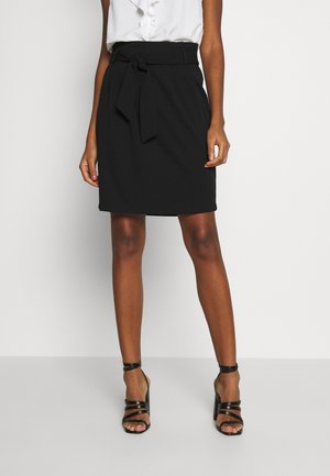 Femme portant une jupe noire longueur genou avec une ceinture nouée à la taille, un chemisier blanc et des sandales noires à talons hauts avec des lanières, debout sur un sol gris.