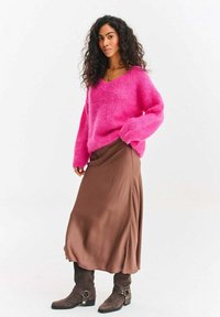 Mäkký ružový sveter s výstrihom do V, hnedá saténová midi sukňa a sivé semišové členkové čižmy so sponami, štýlovo skombinované v stojacej póze.