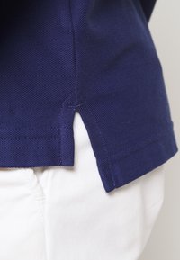 Polo shirt bleu marine texturé avec une fente sur le côté à l'ourlet. La chemise est associée à un pantalon blanc, mettant en avant le contraste des couleurs.